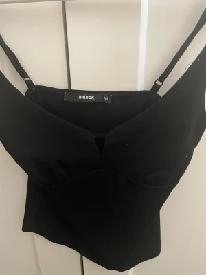 Svart croppad topp från BikBok - Snygg svart croppad topp från BikBok med smala axelband och v-ringad skärning framtill. Perfekt att matcha med höga jeans eller kjol. Enkel och stilren design som passar till många olika looks.