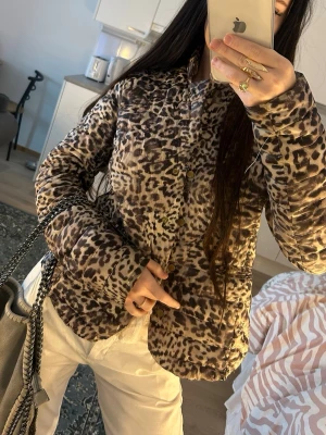 Leopardmönstrad pufferjacka - Säljer en trendig pufferjacka med leopardmönster i beige, brunt och svart. Jackan har tryckknappar framtill, två stora fickor och en lätt vadderad känsla. Perfekt för dig som vill sticka ut och hålla värmen under vintern. Snygg att styla med både jeans och kjol.