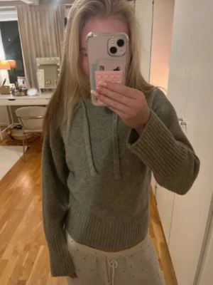 Grå stickad hoodie med huva - Mysig grå stickad hoodie med huva och dragsko. Tröjan har breda ribbade muddar vid ärmslut och midja, vilket ger en avslappnad och trendig look. Perfekt för dig som gillar en chill och stilren vibe.