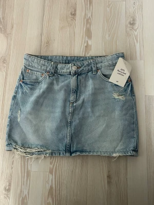 Ljusblå jeanskjol från H&M - Trendig ljusblå jeanskjol från H&M med slitna detaljer och rå kant nertill. Kjolen har klassisk femficksmodell, knapp och dragkedja framtill samt bälteshällor. Perfekt för en avslappnad och cool stil. Storlek 170 men passar mig som har xs/s