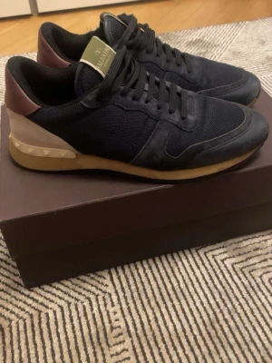 Valentino svarta sneakers med nitar - Snygga svarta sneakers från Valentino med detaljer i mocka och mesh. Skorna har beige och vinröda partier samt ikoniska pyramidnitar på hälen. Yttersulan är i gummi med kontrastfärg. Perfekta för dig som gillar exklusiv streetstyle. Skorna är använda ett fåtal gånger inga större defekter. Perfekta nu när de är vinter. Skriv vid minsta förfrågningar, skorna tvättas innan. Priset är ej hugget i sten.