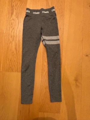 Grå leggings från Stronger i XS - Mörkgrå leggings från Stronger med elastisk midja där loggan STRONGER syns runtom. Byxorna har två vita ränder på ena benet och är i stretchigt material, perfekta för träning. 