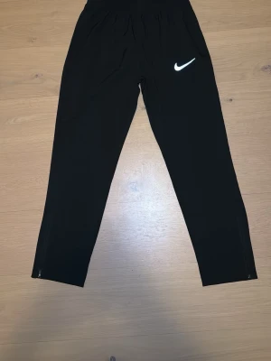 Svarta Nike Windbreaker byxor  - Säljer ett par svarta windbreaker byxor från Nike med klassisk vit logga på låret. Byxorna har dragkedjor vid bensluten och elastisk midja för skön passform. Perfekta för löpning eller gym. Materialet känns lätt och smidigt. Storleken är XL men passar som L