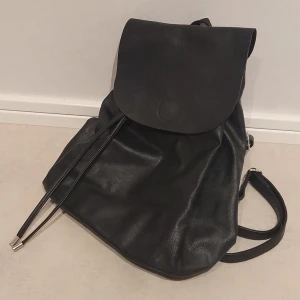 Svart ryggsäck i läderlook - Stilren svart ryggsäck med slät läderlook och enkel design. Ryggsäcken har ett stort lock med magnetknäppning och justerbara axelband. Perfekt för dig som vill ha en minimalistisk och snygg accessoar till skolan eller stan.