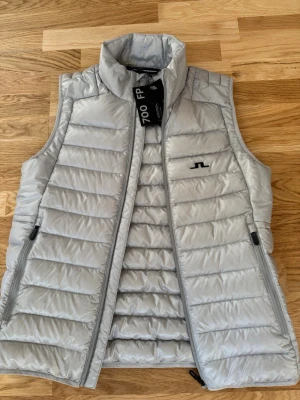 Ljusgrå dunväst från J.Lindeberg - Snygg ljusgrå dunväst från J.Lindeberg med hög krage och två dragkedjeförsedda fickor. Västen har quiltad design och diskret logga på bröstet. Tillverkad i lätt material med 700 fill power dun för extra värme. Perfekt för lager-på-lager-stil. Original pris ca3500kr