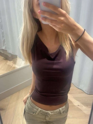 Mörkbrun ärmlös topp från Gina Tricot - Säljer en mörkbrun ärmlös topp från Gina Tricot med snygg draperad halsringning. Toppen är croppad och har en tight passform som framhäver figuren. Perfekt att styla med jeans eller kjol för en trendig look.💞💞