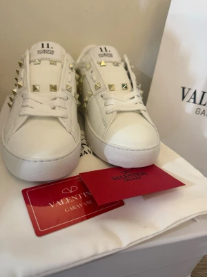Vita Valentino Garavani sneakers - Snygga vita sneakers från Valentino Garavani med ikoniska guldfärgade nitar längs sidorna och på plösen. Skorna är i läder med klassisk låg siluett och vita skosnören. Perfekta för dig som vill sticka ut med en exklusiv touch.