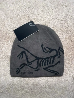 Grå mössa från Arc'teryx - Snygg grå mössa från Arc'teryx med deras ikoniska fossil-logga i svart. Tillverkad i mjukt, stickat material som sitter skönt på huvudet. Perfekt för dig som gillar stilrena accessoarer med en sportig touch. Perfekt nu under kallt väder!❄️
