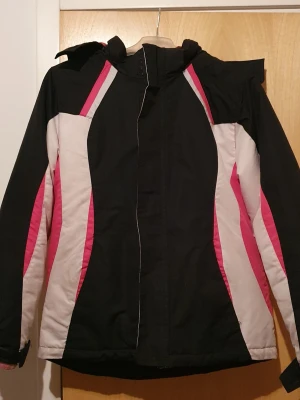 Vinterjacka - Snygg vinterjacka fodrad i svart med vita och rosa detaljer. Jackan har huva, dragkedja framtill och sportig design med färgblock längs sidorna och ärmarna. Perfekt för kalla dagar och har en modern look som sticker ut. Det är i storlek M i barnstorlek. Finns flera fläckar.