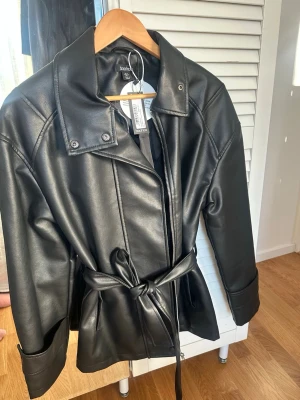 Svart skinnjacka från Boohoo - Snygg svart skinnjacka från Boohoo med bälte i midjan och bred krage. Passar S också