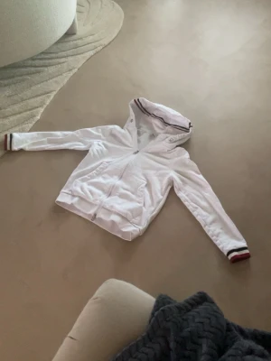 Vit hoodie från Moncler med ränder - Snygg vit hoodie från Moncler med dragkedja framtill och huva. Detaljer i form av röda och svarta ränder på muddar och huva samt Moncler-logga på bröstet. Tillverkad i mjukt bomullsmaterial och har två fickor framtill.