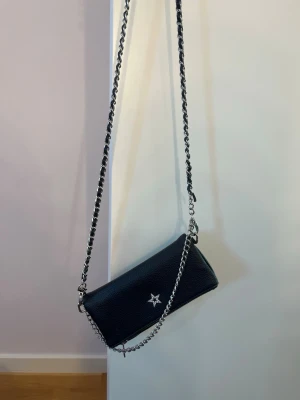 Svart axelväska - Svart handväska med en stjärna på framsidan och en liten kedja. På den långa kedjan man hänger väskan i, har remmen gått ut ur kedjan lite men det går nog att laga om man pillar lite. Väskan har använts många gånger men det syns inte. Den ser nästan helt ny ut. Den är från Gina tricot. 