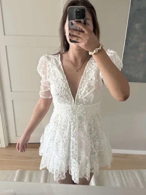 Studentklänning  - Superfin vit kort klänning med djup v-ringning och puffärmar. Klänningen har spetsdetaljer och blommigt broderi över hela tyget. Perfekt för sommarens tillfällen och student. Luftigt material och markerad midja ger en söt siluett.