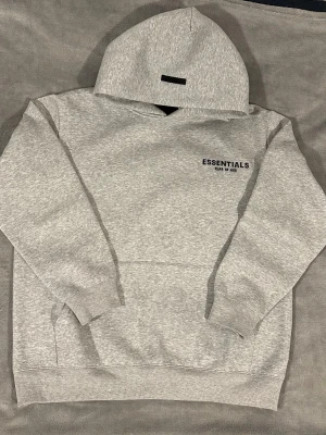 Essentials Fear of God hoodie - Säljer en grå hoodie från Essentials Fear of God med svart logga på bröstet och stor text på ryggen. Hoodien har en klassisk huva, ribbade muddar och magficka. Tillverkad i mjukt bomullsmaterial som är perfekt för chill dagar.
