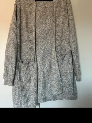 Grå stickad kofta med huva - Mysig gråmelerad stickad kofta med huva och två stora fickor framtill. Koftan har lång ärm och är öppen framtill utan knäppning. Perfekt för lager-på-lager och chill dagar. Materialet är grovstickat och känns riktigt skönt.