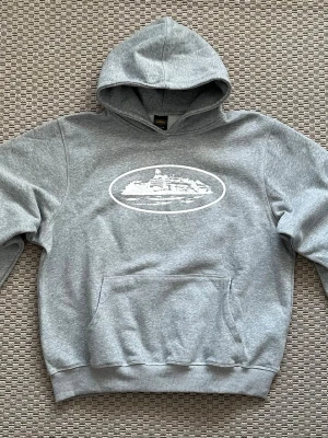 Corteiz hoodie  - Snygg grå hoodie från Corteiz med vit tryckt grafik på bröstet som föreställer en byggnad i en oval ram. Klassisk känguruficka framtill och ribbade muddar vid ärmar och nederkant. Perfekt för en avslappnad streetwear-look.