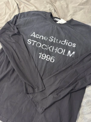 Svart långärmad topp Acne Studios - Svart långärmad topp från Acne Studios med tryck 'Acne Studios STOCKHOLM 1996' framtill. Klassisk rund halsringning och relaxed fit. Perfekt för dig som gillar stilrena och trendiga plagg med tydlig logga.