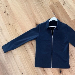 Mörkblå overshirt från Jack & Jones - Stilren mörkblå overshirt från Jack & Jones med diskret struktur i tyget. Modellen har två diskreta bröstfickor, klassisk krage och hel dragkedja framtill. Perfekt lager på lager plagg med snygg passform. Medeltjockt material.