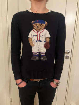 Svart Polo Ralph Lauren tröja stickad - Svart stickad tröja från Polo Ralph Lauren med ikoniska Polo Bear i basebolloutfit på bröstet. Rund halsringning och ribbstickade muddar vid ärmslut och nederkant. Perfekt för dig som gillar sportig och lekfull stil. Bara att höra av sig vid fundering!!