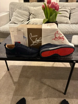 Marinblå sneakers från Louboutin - Snygga marinblå sneakers från Christian Louboutin med klassisk röd sula och diskreta svarta detaljer. Skorna har lågt skaft, svarta skosnören och ovandel i textil och läder. Perfekta för dig som vill ha en exklusiv och stilren look. Små defekter på höger skon som visas på bild 4/5 men lätt att fixa. Har haft dom ett tag men endast använt dom ett fåtal gånger därav skicket, skulle säga att dom är 8-9/10 om 10 är helt nya, kontakta vid frågor. Pruttat och klart