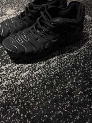 Nike Air VaporMax Plus svarta sneakers - Svarta Nike Air VaporMax Plus sneakers med textil- och syntetmaterial, ikoniska vågformade detaljer på ovandelen och synlig VaporMax-sula. Klassisk snörning och diskret logga på hälen. Riktigt stilrena och passar perfekt till streetwear.
