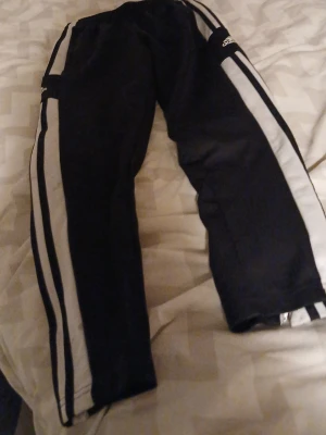 Svarta Adidas träningsbyxor - Säljer ett par svarta träningsbyxor från Adidas med vita ränder längs sidorna och Adidas-logga på benet. Byxorna har en klassisk sportig look och är perfekta för gymmet eller fritiden. Tillverkade i ett lätt och bekvämt material.