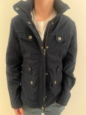 Mörkblå jacka från Sttaccato 152cm - Snygg mörkblå jacka från stacco med hög krage, dragkedja och tryckknappar. Jackan har fyra fickor framtill med lock och knapp, samt detaljer i metall. Materialet är slitstarkt och jackan har en sportig, avslappnad look.