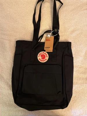 Svart Kånken ryggsäck från Fjällräven - Klassisk svart Kånken ryggsäck från Fjällräven med det ikoniska runda röda logotypmärket framtill. Ryggsäcken har en stor ytterficka med dragkedja, två sidofickor och långa bärhandtag. Tillverkad i slitstarkt syntetmaterial.