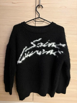 Svart stickad YSL tröja  - Säljer en svart stickad tröja med vit text i grafisk stil över bröstet. Tröjan har lång ärm, rund halsringning och är gjord i ett fluffigt material som känns riktigt mysigt. Perfekt för dig som gillar en avslappnad och trendig look.