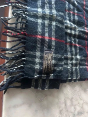 Mörkblå rutig halsduk från Burberry - Klassisk halsduk från Burberry i mörkblått med vitt och rött rutmönster. Halsduken har fransar i ändarna och är tillverkad i 100% lammull, vilket gör den både mjuk och varm. Perfekt accessoar för att lyfta din outfit med en tidlös look.