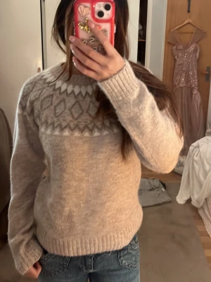 Beige stickad tröja med mönster - Mysig beige stickad tröja med rund hals och ett snyggt mönster i vitt och grått över bröstet och axlarna. Tröjan har ribbade muddar vid ärmslut och nederkant, perfekt för kalla dagar. Passar till jeans eller kjol för en chill look.