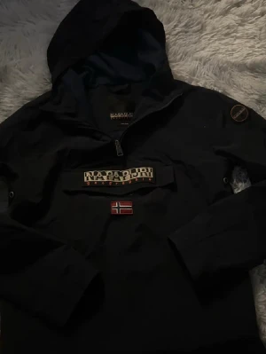 Svart vindjacka från Napapijri - Svart vindjacka från Napapijri med huva och halv dragkedja framtill. Stor logotyp på bröstet och norsk flagga under. Jackan har en ficka framtill och patch på ärmen. Perfekt för blåsiga dagar och streetwear-stil.