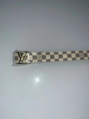 Bälte - Snyggt bälte från L med klassiskt rutigt mönster i beige och brunt. Bältet har ett stort guldfärgat L-spänne och är tillverkat i skinn. Perfekt accessoar för att lyfta din outfit.