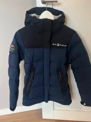 Mörkblå dunjacka Sail Racing - Snygg mörkblå dunjacka från Sail Racing med svart axelparti och huva. Jackan har två dragkedjeförsedda fickor fram, logga på bröstet och ärmen samt quiltad design. Perfekt för kalla vinterdagar och riktigt skön passform.