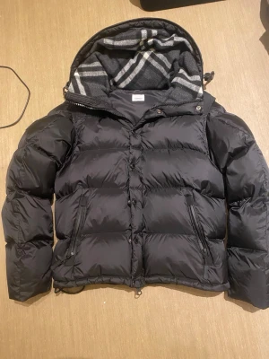 Burberry jacka - Stilren svart pufferjacka från Burberry med stor huva och klassiskt rutigt foder. Jackan har dragkedja, knappar och två snedställda fickor med dragkedja. Tydlig Burberry-logga på ärmen. Perfekt för kalla vinterdagar.