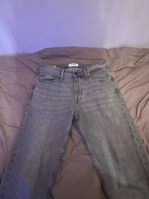 Grå raka jeans från Jack&Jones - Säljer ett par grå jeans från ARKET med rak passform och klassisk femficksdesign. Jeansen har hög midja, bälteshällor och är gjorda i kraftig denim. Snyggt minimalistiskt läderpatch bak i midjan och diskreta sömmar. Perfekta till en avslappnad look.
