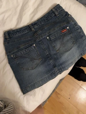 Blå vintage jeanskjol - Säljer en blå vintage jeanskjol i kort modell. Kjolen har snygga bakfickor med dekorativa sömmar och en liten röd detalj på ena fickan. Perfekt till vardags och enkel att styla med olika toppar. 