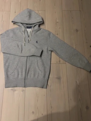 Grå zip hoodie från Polo Ralph Lauren - Snygg gråmelerad hoodie från Polo Ralph Lauren med dragkedja, två fickor fram och klassisk huva med vita snören. Liten mörkblå logga broderad på bröstet. 