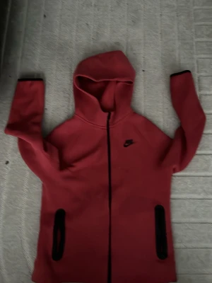 Röd Nike hoodie med dragkedja - Säljer en röd hoodie från Nike med hel dragkedja framtill och svarta detaljer på fickorna. Hoodien har en klassisk huva och Nike-logga på bröstet. Perfekt för dig som gillar sportig och avslappnad stil.