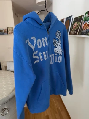 Blå Von Studio hoodie med tryck - Blå hoodie från Von Studio med dragkedja, stor huva och fickor framtill. Framsidan har ett stort vitt tryck med texten 'Von Studio' och en tecknad ängel. Perfekt för dig som gillar streetwear och vill sticka ut med en cool design.