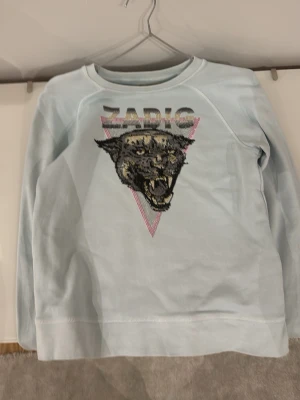 Ljusblå sweatshirt från Zadig - Cool ljusblå sweatshirt från Zadig med ett stort paljettmönster av ett panterhuvud och texten 'ZADIG' på bröstet. Tröjan har rund halsringning och långa ärmar. Perfekt för dig som gillar statement-plagg och djurmotiv.