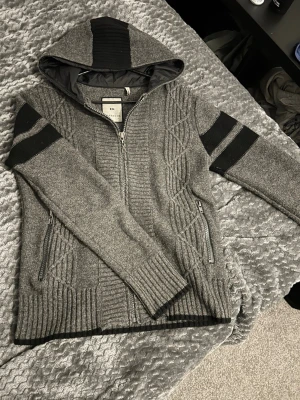 Grå ziphoodie från Freesoul - Grå stickad ziphoodie från Freesoul med svarta detaljer på ärmar och huva. Tröjan har dragkedja framtill, två fickor med dragkedja och ribbstickade muddar. Snyggt mönster längs sidorna och fodrad huva för extra komfort.