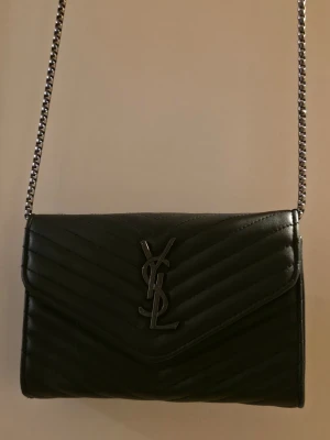 Svart YSL kuvertväska i läder - Snygg svart kuvertväska från Saint Laurent med klassiskt YSL-emblem i metall framtill. Väskan är tillverkad i quiltat läder med chevronmönster och har en silverfärgad kedjerem. Perfekt för att lyfta vilken outfit som helst.