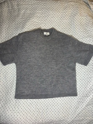 Grå stickad t-shirt från Gina - Säljer en stilren grå stickad t-shirt från Gina . Tröjan har korta ärmar, rund halsringning och ribbade kanter. Materialet är mjukt och ger en avslappnad men ändå snygg look. Perfekt att matcha med jeans eller kjol.