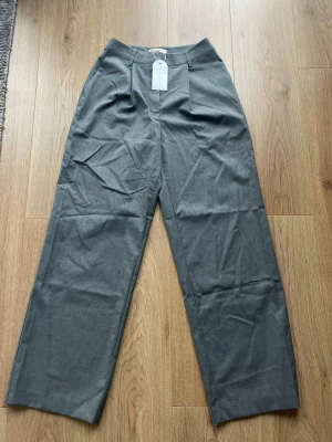 Grå kostymbyxor från Sanne Alexandra - Snygga grå kostymbyxor från Sanne Alexandra med raka ben och hög midja. Byxorna har pressveck framtill, bälteshällor och stängs med dragkedja och hake. Perfekta för en clean och stilren look. Helt nya med lapp kvar!