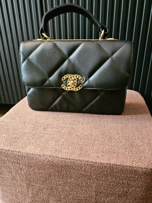 Svart quiltad handväska från C h a n e l - Snygg svart handväska från C h a n e l i quiltat läder med guldfärgade detaljer och ikonisk CC-logga framtill. Väskan har ett topphandtag och elegant, stilren form som ger en lyxig känsla. Perfekt accessoar för dig som vill sticka ut med klassisk design.