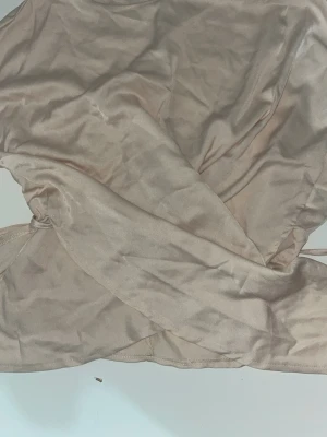 Beige omlottopp i satinlook - Snygg beige omlottopp i glansigt satinliknande material. Toppen har knytband i midjan för en justerbar passform och ger en elegant känsla. Perfekt att styla med jeans eller kjol för en trendig look.