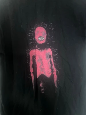 Svart t-shirt med röd skräckgrafik - Säljer en svart t-shirt med en unik och edgy röd grafik framtill som föreställer en skräckinspirerad figur med synliga tänder och elektriska linjer runt huvudet. T-shirten har korta ärmar och är gjord i mjuk bomull. Perfekt för dig som gillar alternativ stil.