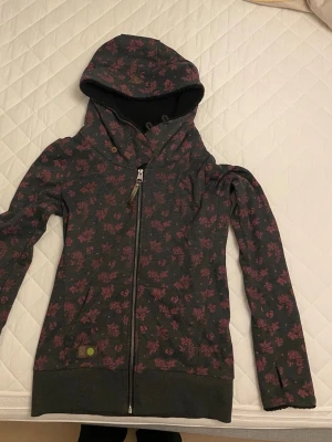 Mörkgrå hoodie med rosa blommor Ragwear - Mysig mörkgrå hoodie från Ragwear med rosa blommönster och dragkedja framtill. Hoodien har en stor huva med mjukt, svart foder och praktiska fickor. Tillverkad i ekologiskt material och har tumhål vid ärmsluten för extra komfort.