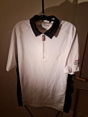 Vit och svart polotopp Oscar Jacobson - Vit polotopp från Oscar Jacobson med svarta detaljer på krage och sidor. Halv dragkedja framtill och korta ärmar. Broderad logga på ärmen. Perfekt för en sportig och stilren look.